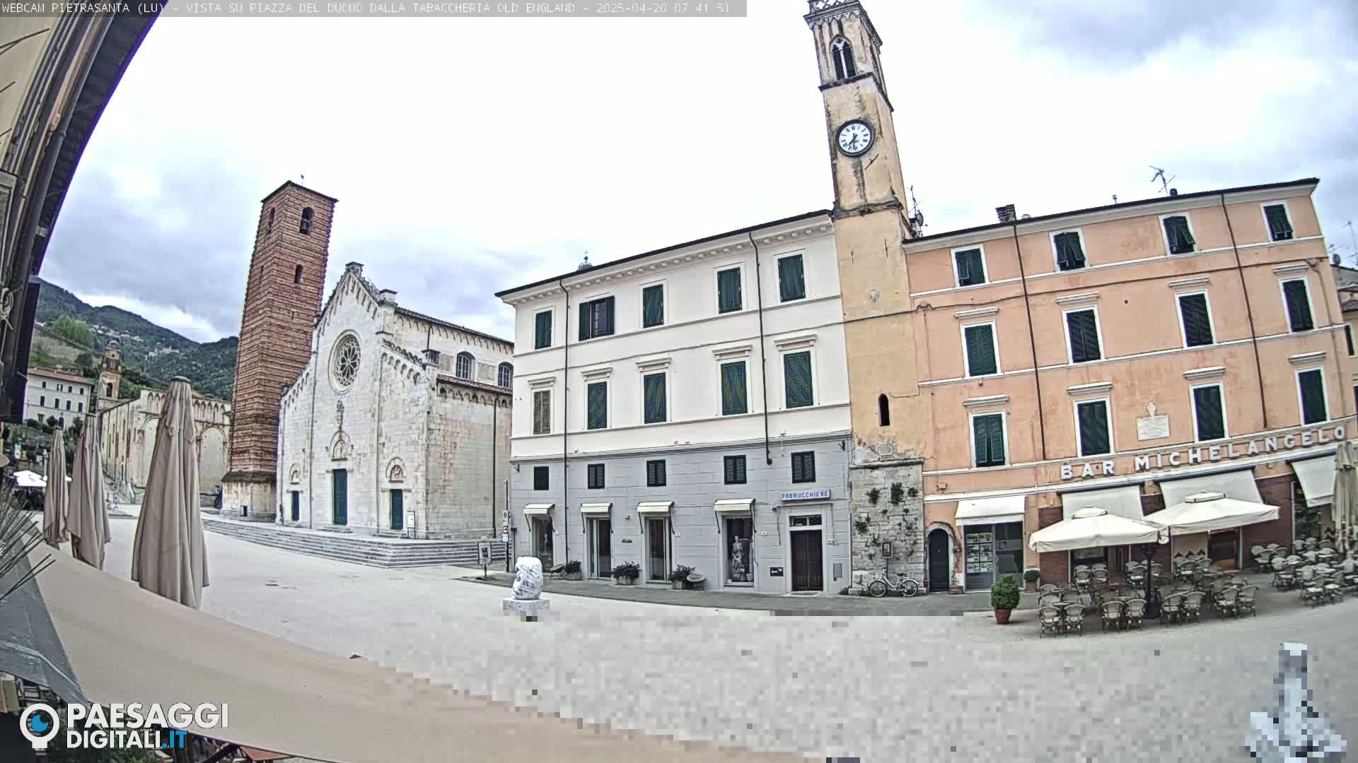 A town square with several buildings, including a church with a tall bell tower and a peach-colored building with outdoor seating, under an overcast sky.