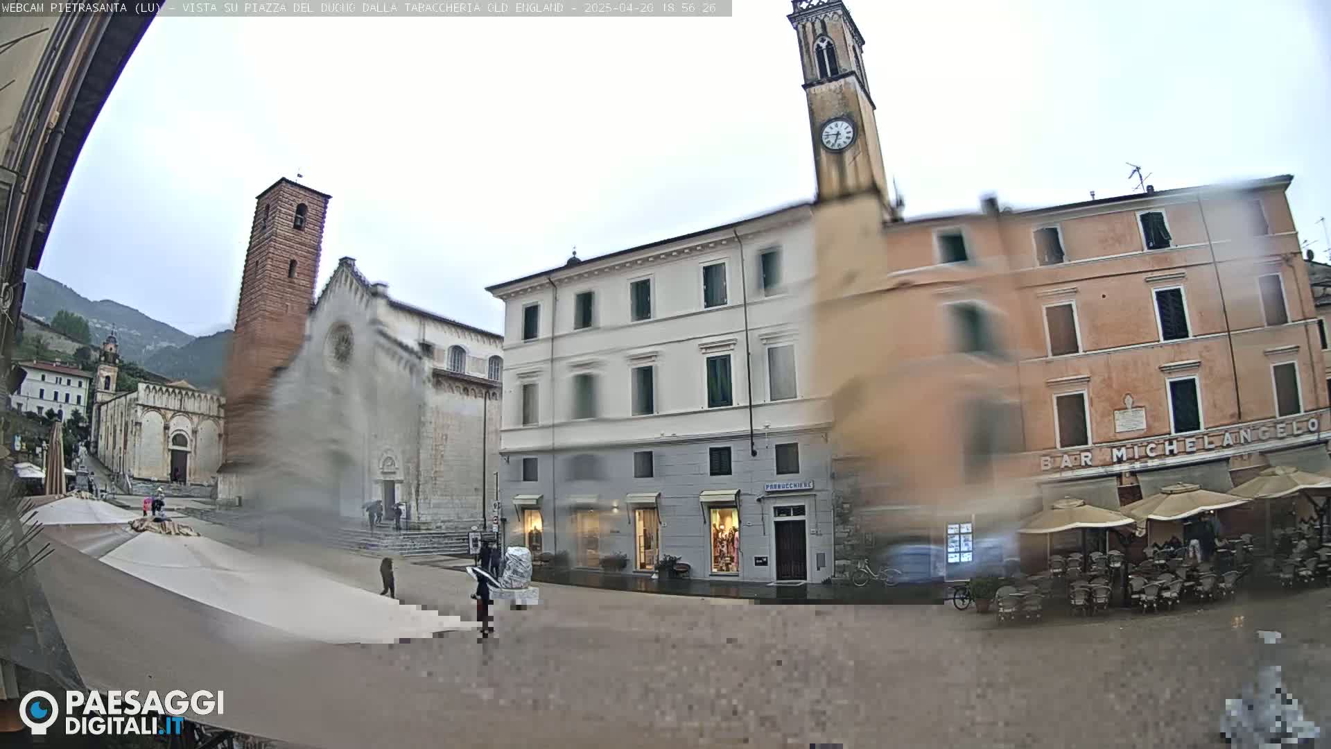 A rain-slicked town square in Italy, featuring several buildings, a church with a tall bell tower, and outdoor café seating.