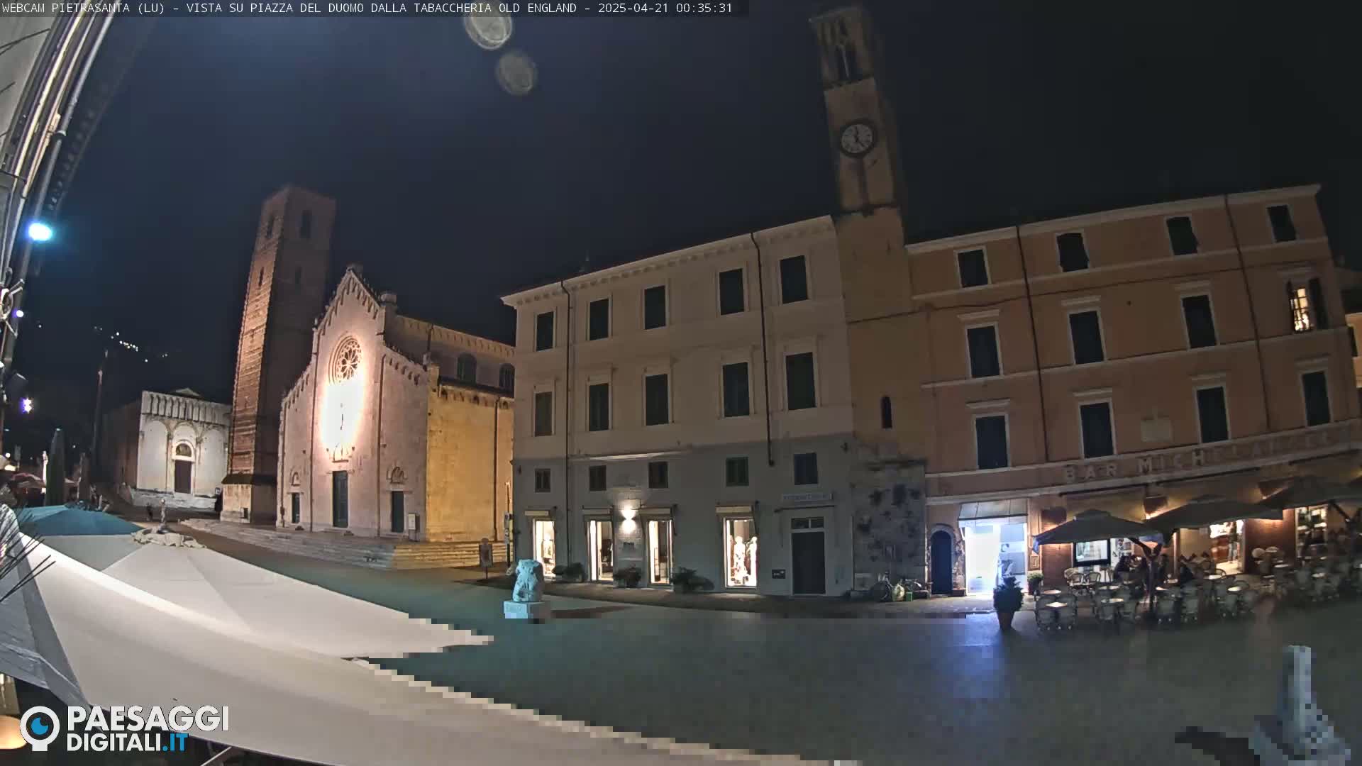 A nighttime view of a town square with a church, buildings, and an outdoor cafe under a clear sky.