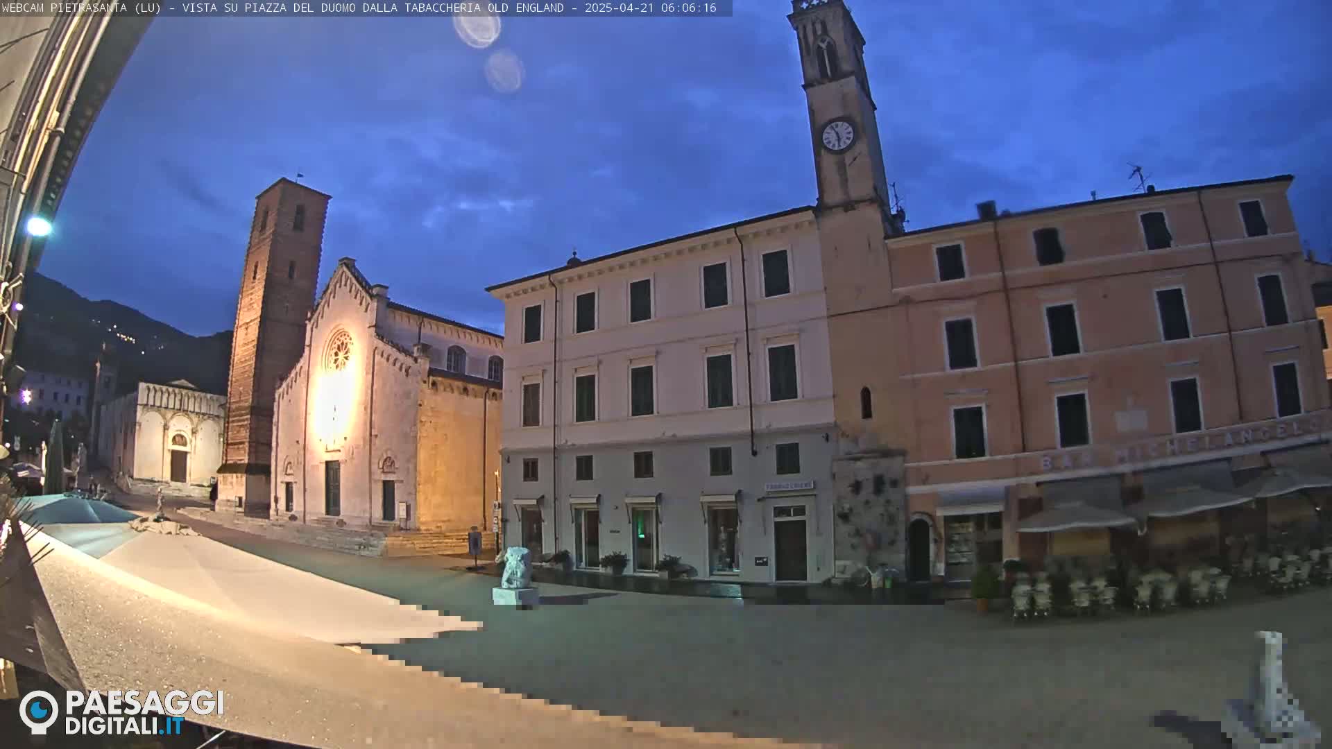 A town square at twilight, showing a church with a tall bell tower, other buildings, and a mostly empty square.