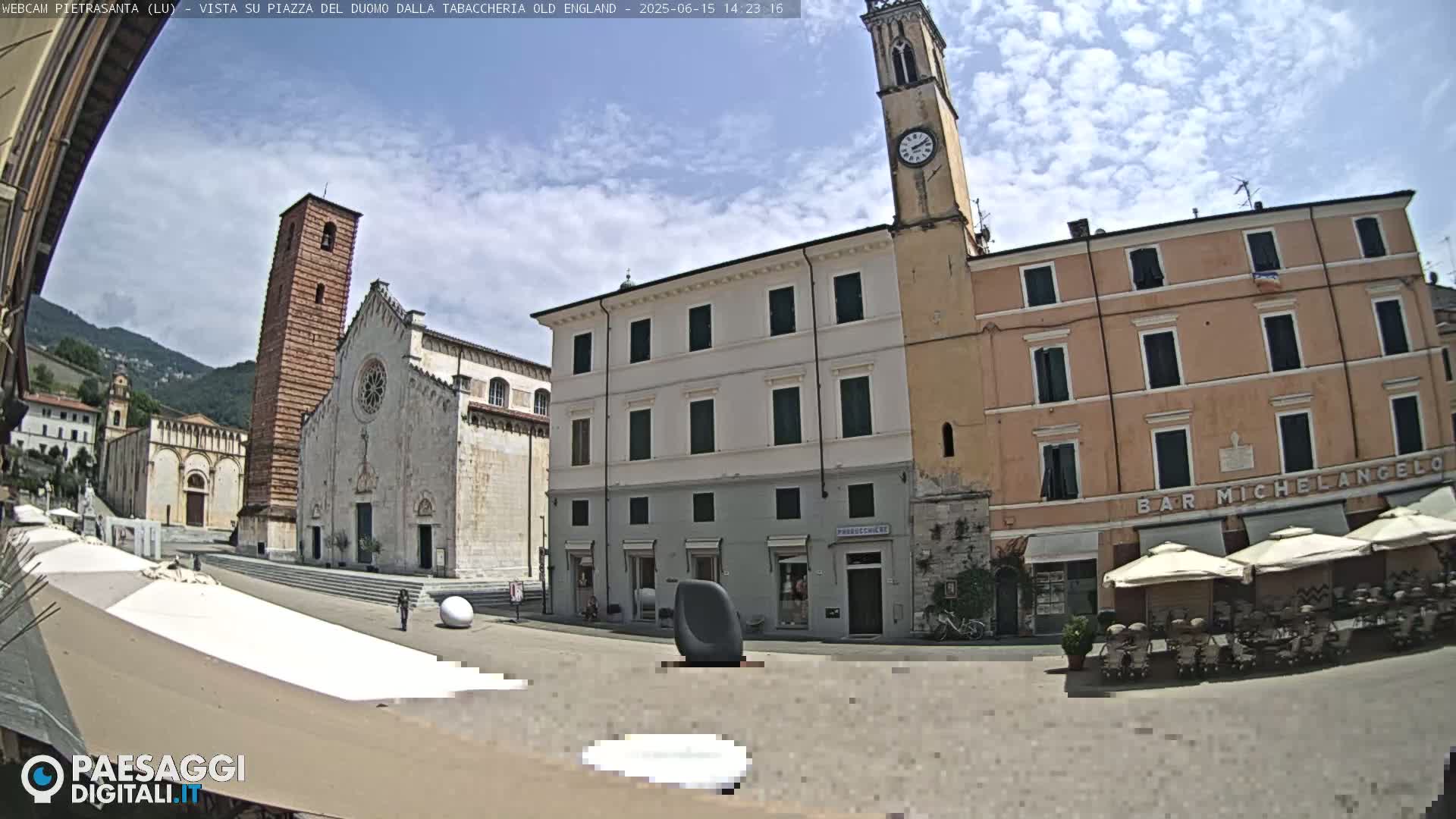 A town square is shown on a partly cloudy day, with a large church, several buildings, and outdoor seating.
