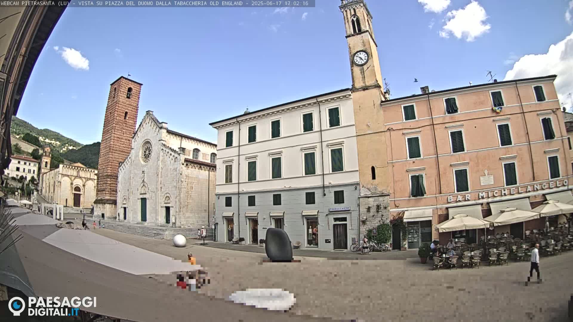 A sunny town square in Italy, featuring a large cathedral, surrounding buildings, a clock tower, and an outdoor cafe with patrons sitting at tables under umbrellas.