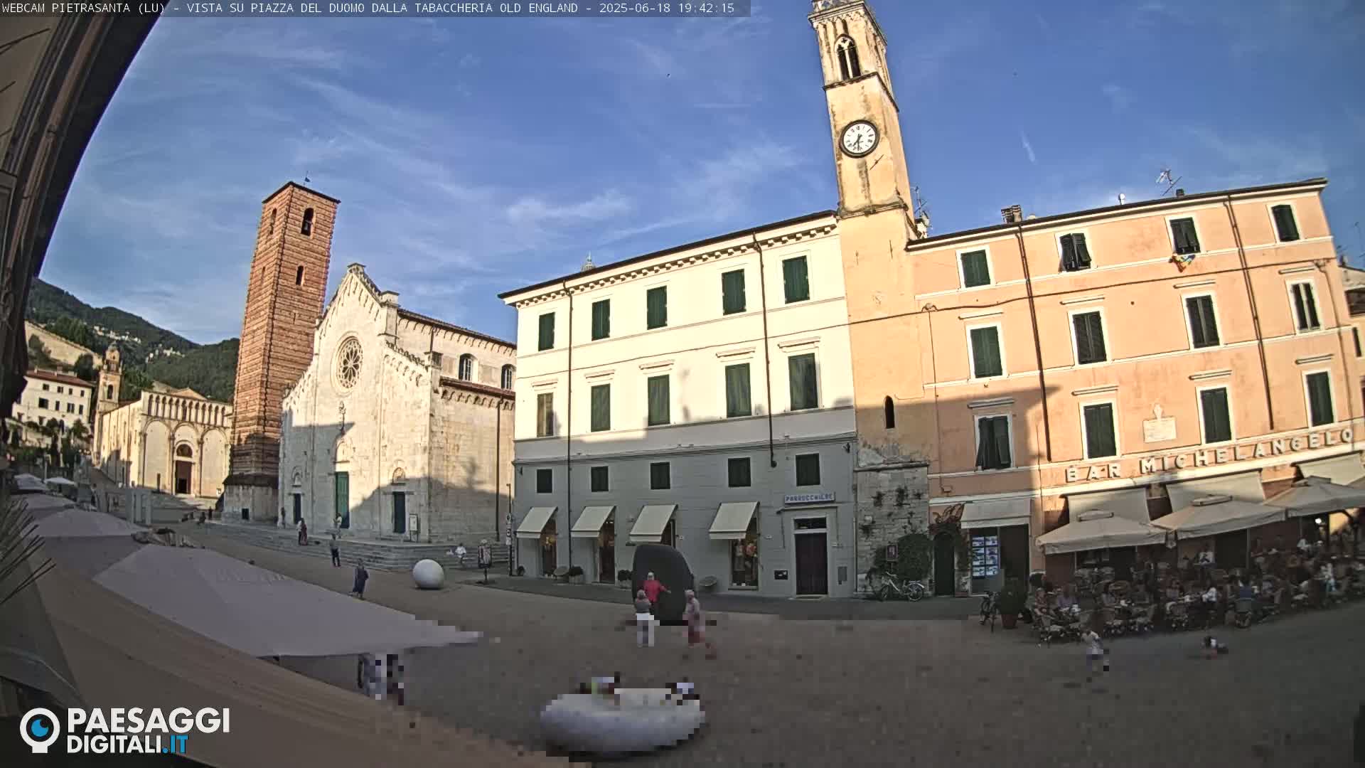 A sunny town square in Italy features a large cathedral, a clock tower, and several buildings, with people sitting at outdoor cafes under umbrellas.