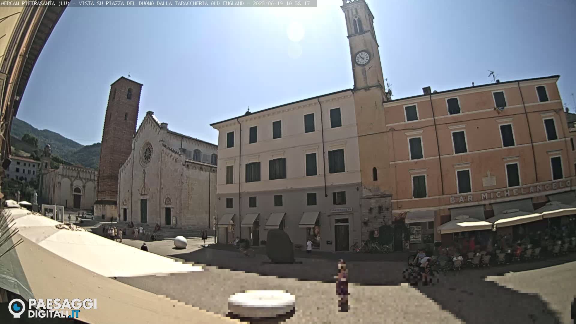 A sunny town square is shown, featuring a large cathedral, adjacent buildings, and people sitting at outdoor cafes.
