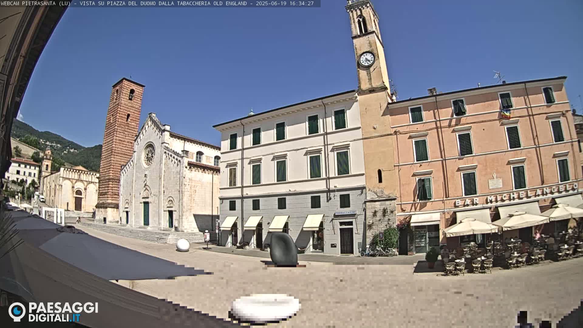 A sunny town square is shown with a large church, clock tower, and buildings, with outdoor seating at a cafe.