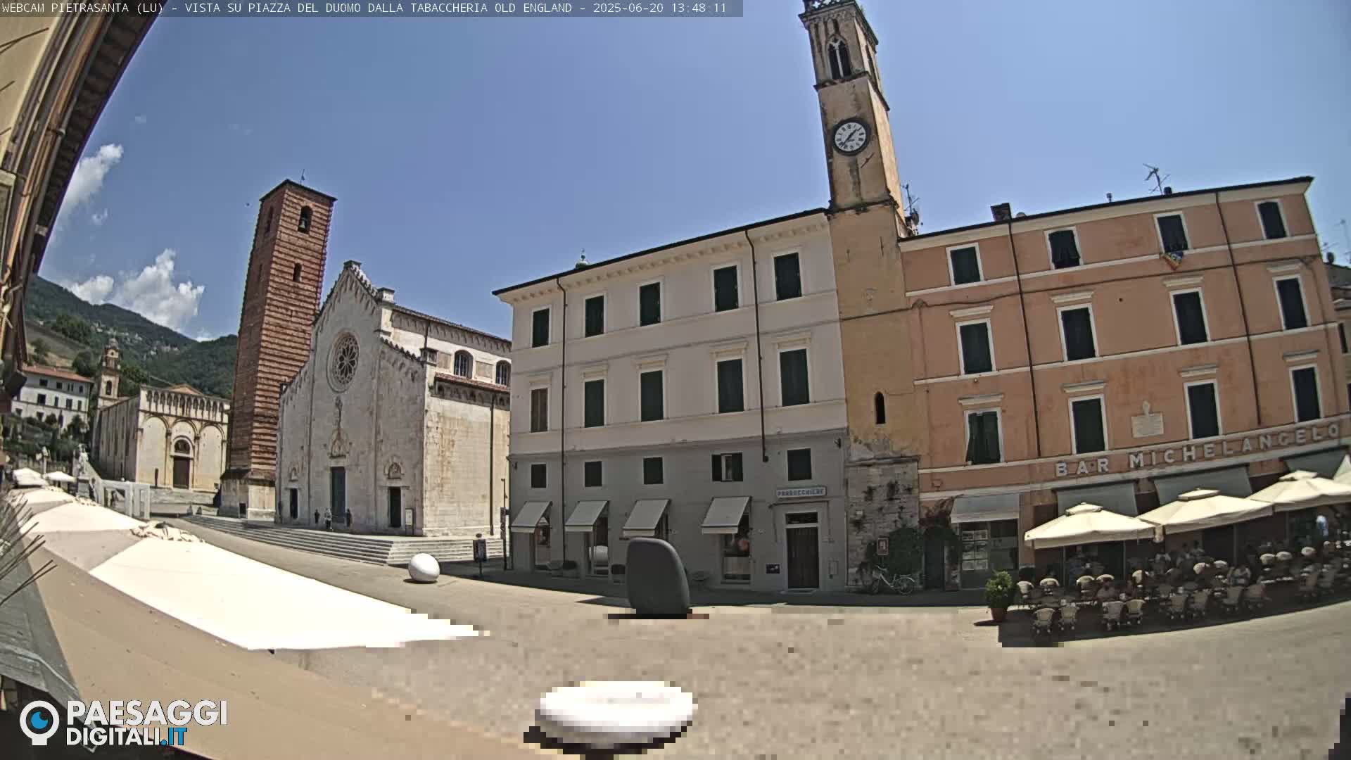 A sunny town square in Italy features a large cathedral, a clock tower, several buildings, and outdoor seating at a café, all under a clear blue sky.