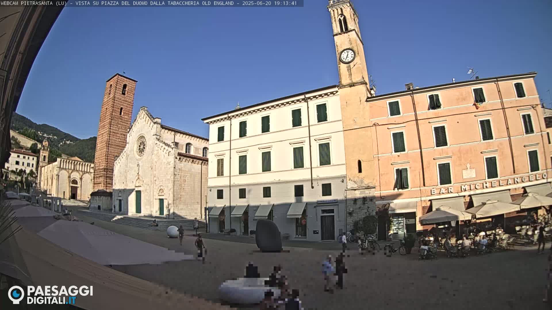 A sunlit town square in Italy, featuring a cathedral, a clock tower, and several buildings, with people sitting at outdoor cafes under umbrellas.