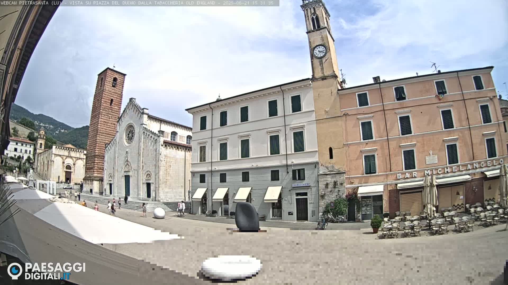 A sunny town square with a large church, several buildings, a clock tower, outdoor seating, and a few people.