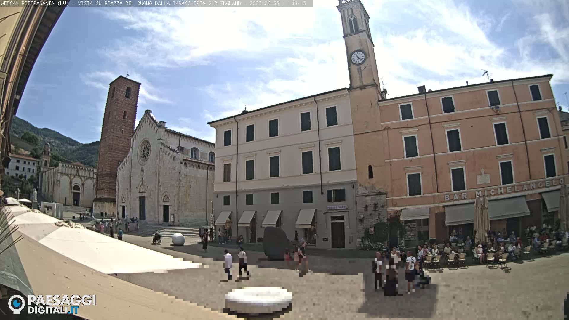 A sunny town square with a large cathedral and clock tower, surrounded by buildings and people sitting at outdoor cafes.