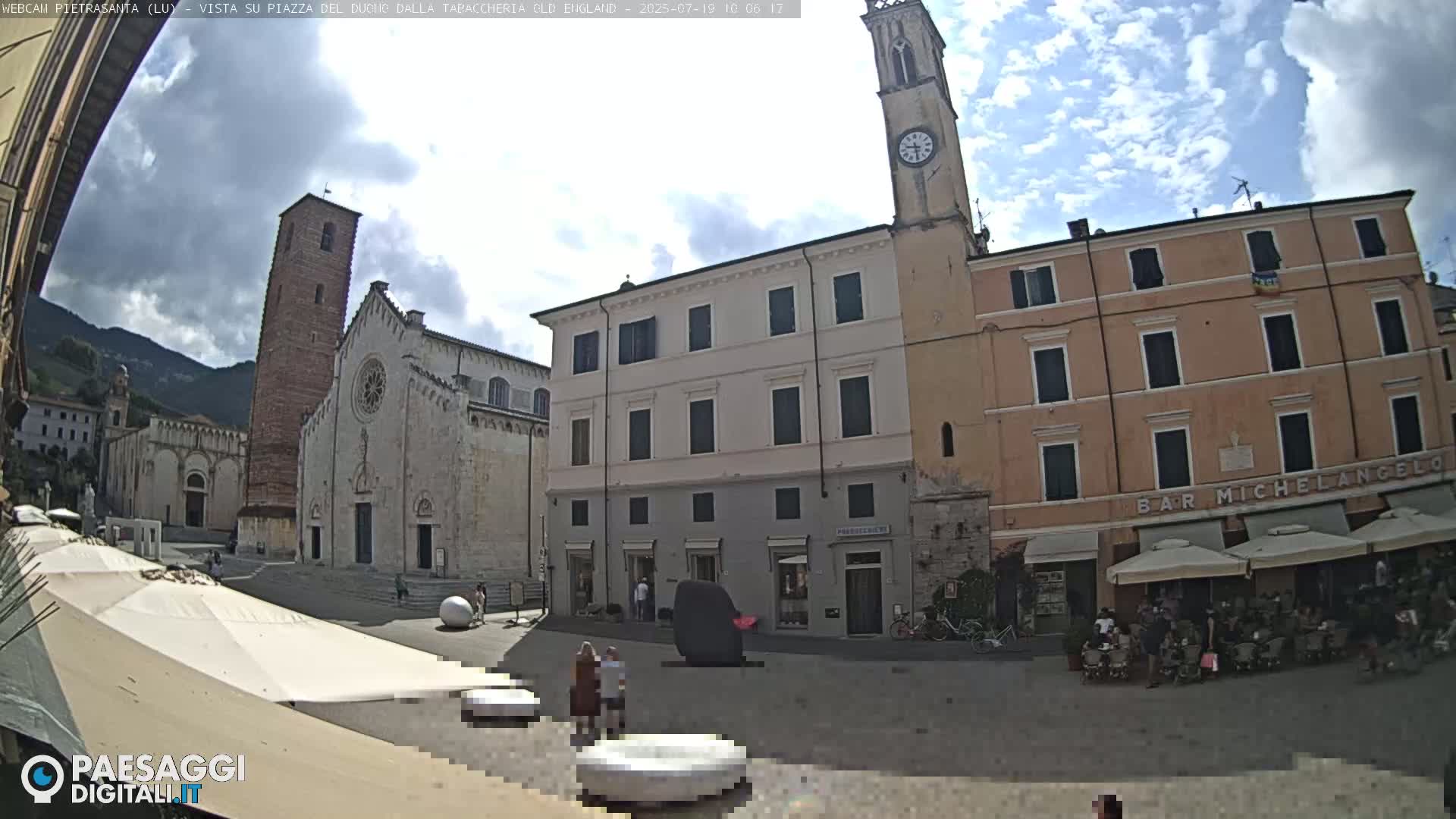 A partly cloudy day in a town square with several buildings, including a cathedral and clock tower, and outdoor seating at a cafe.