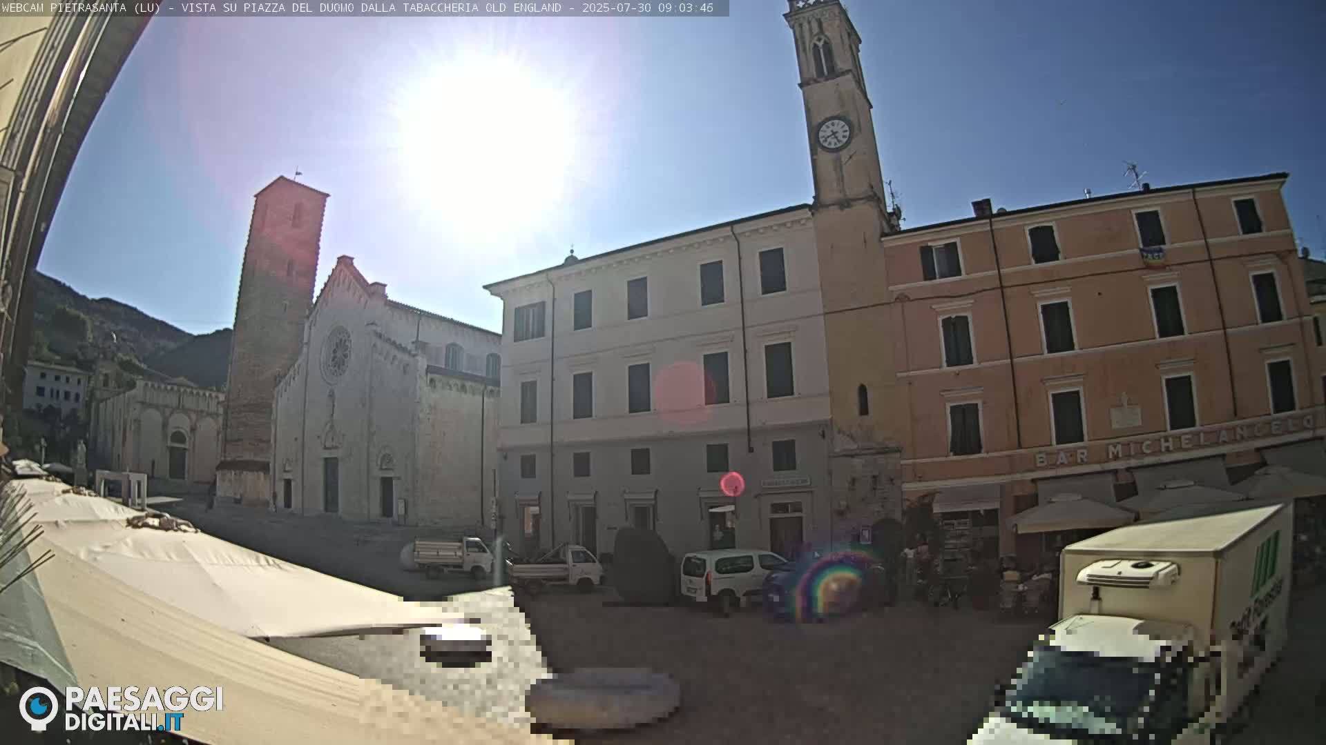 A sunny town square with a large church, several buildings, and vehicles parked.