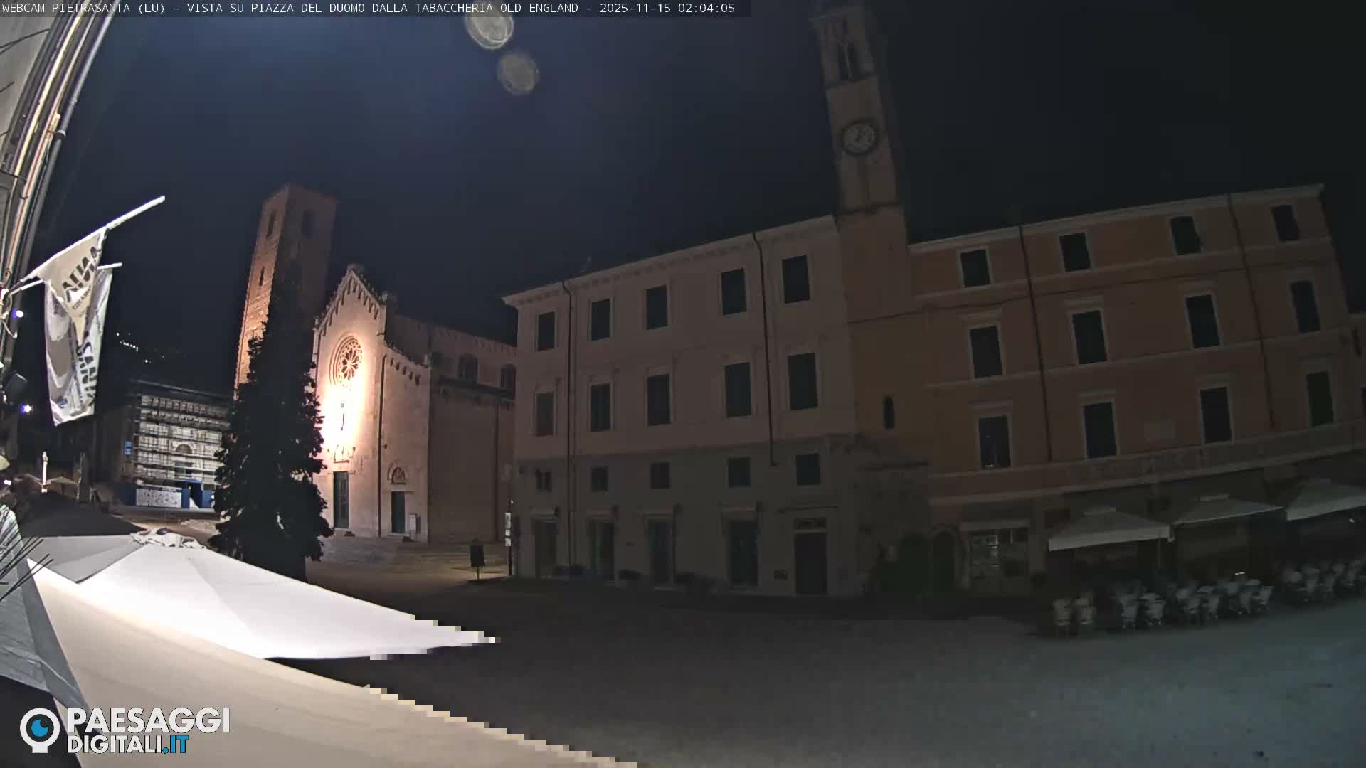 A clear night illuminates a European town square, featuring a brightly lit church with a tall bell tower, historical buildings including one with a clock tower, outdoor cafe seating, and a lone pedestrian.
