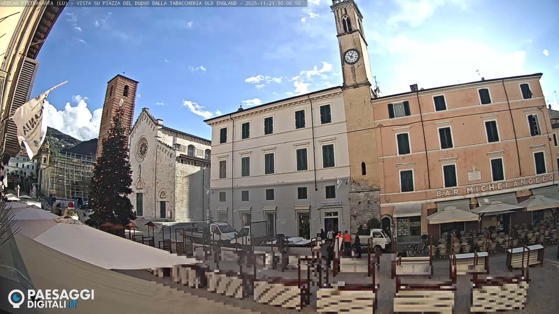 A sunny day reveals a historic European town square featuring an ancient church with a tall bell tower and a prominent Christmas tree, surrounded by multi-story buildings and an outdoor café area with tables and chairs.