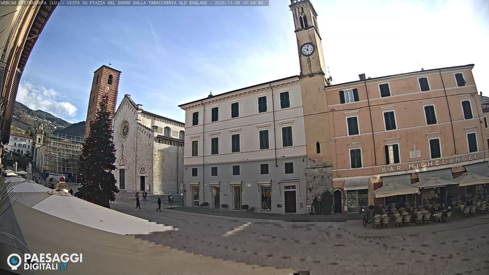 A sunlit European town square showcases a large stone cathedral and a prominent brick clock tower alongside a decorated Christmas tree, with multi-story buildings and an outdoor cafe, all beneath a clear blue sky framed by distant mountains.