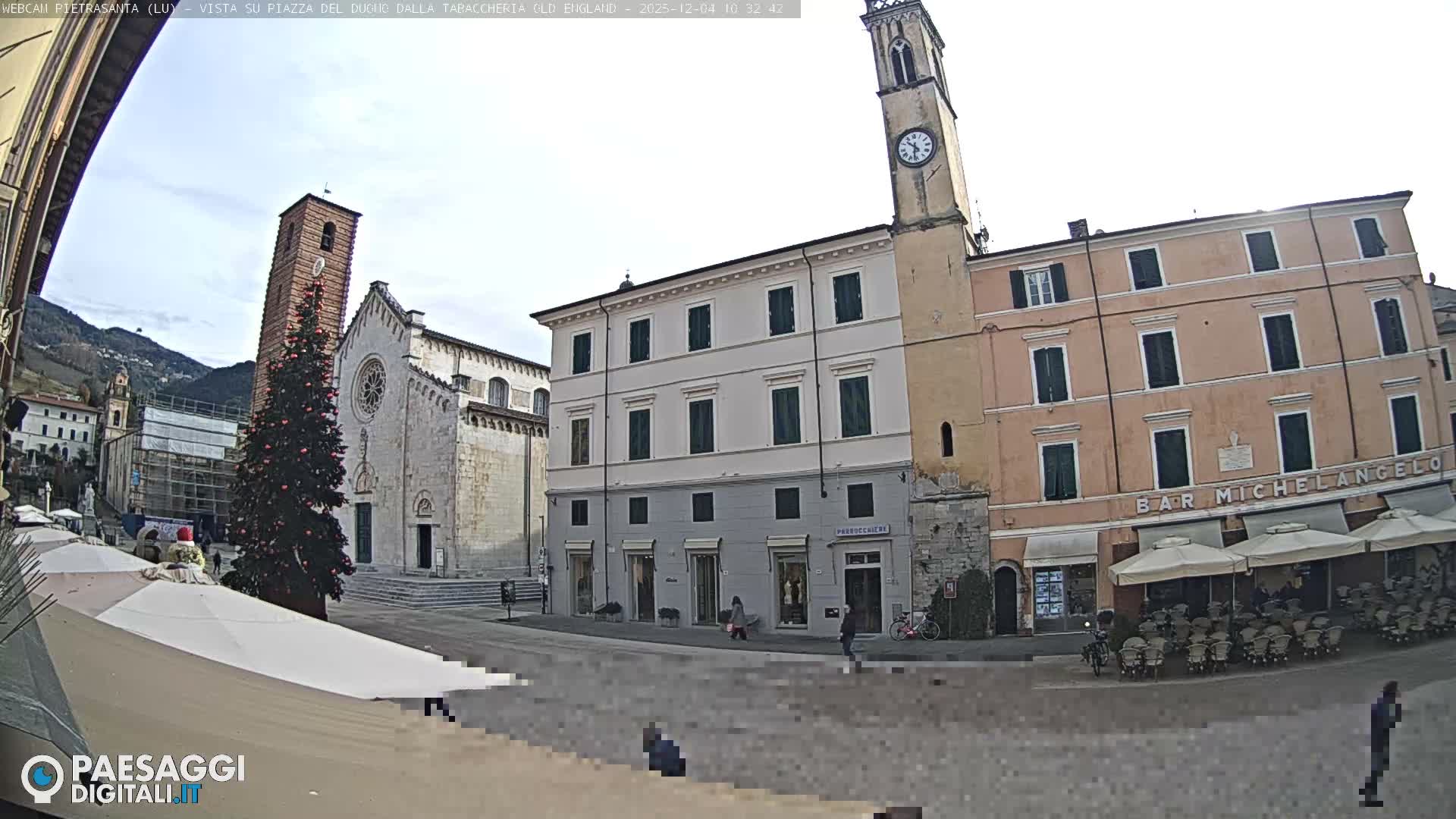 A wide view of an Italian town square on an overcast day features a large Christmas tree next to a historic church and bell tower, surrounded by traditional multi-story buildings and a cafe with outdoor seating.