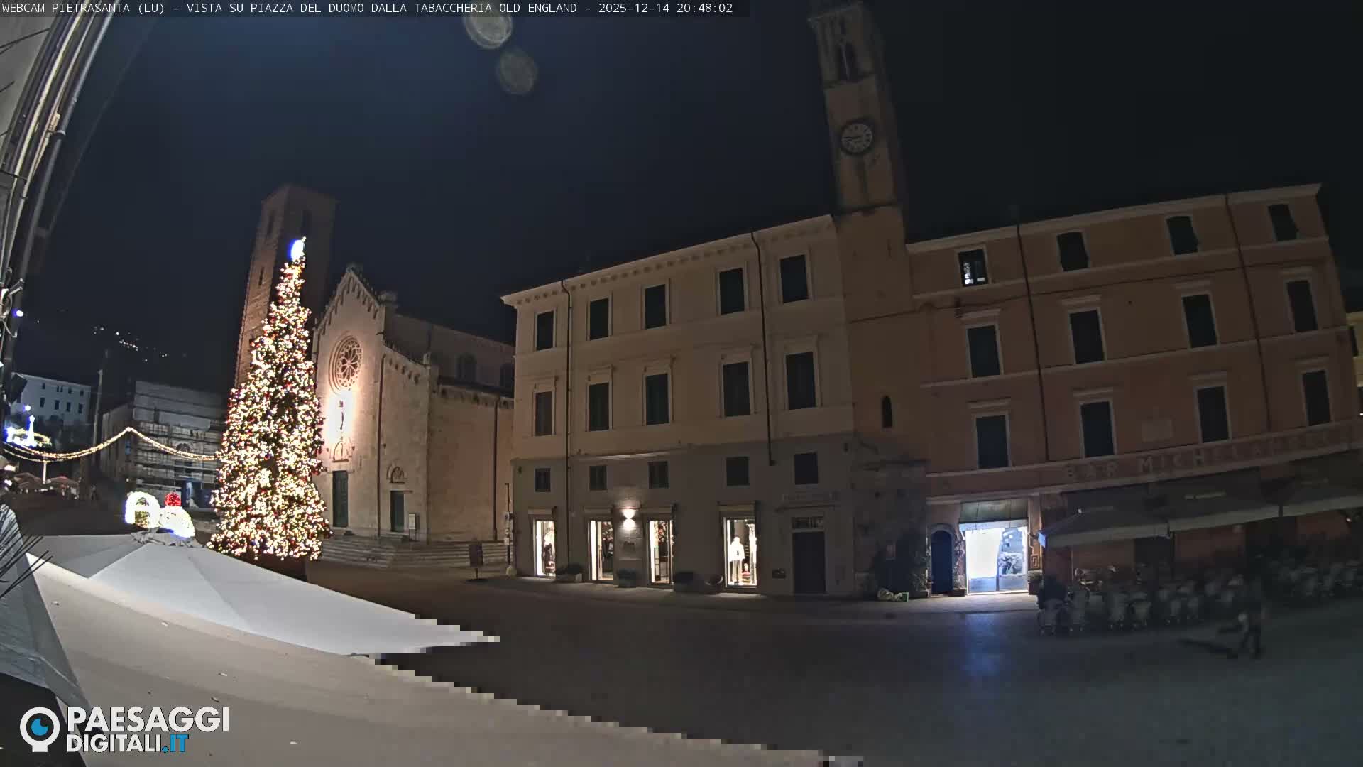 A wide view of an Italian town square on an overcast day features a large Christmas tree next to a historic church and bell tower, surrounded by traditional multi-story buildings and a cafe with outdoor seating.