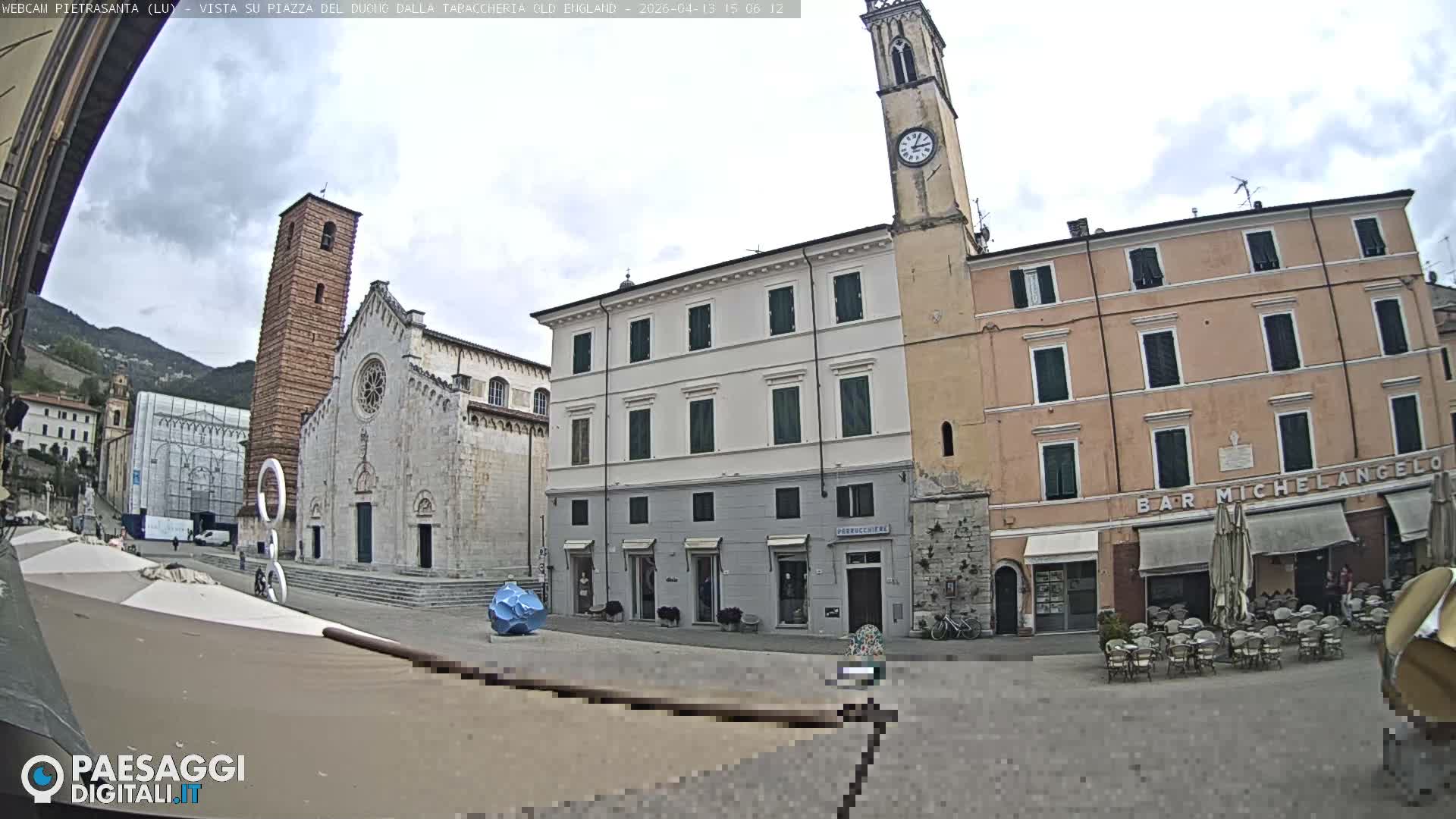 A wide view of an Italian town square on an overcast day features a large Christmas tree next to a historic church and bell tower, surrounded by traditional multi-story buildings and a cafe with outdoor seating.