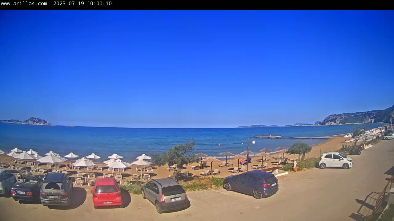 A sandy beach with numerous umbrellas and lounge chairs is visible, with several cars parked nearby, all under a clear blue sky.
