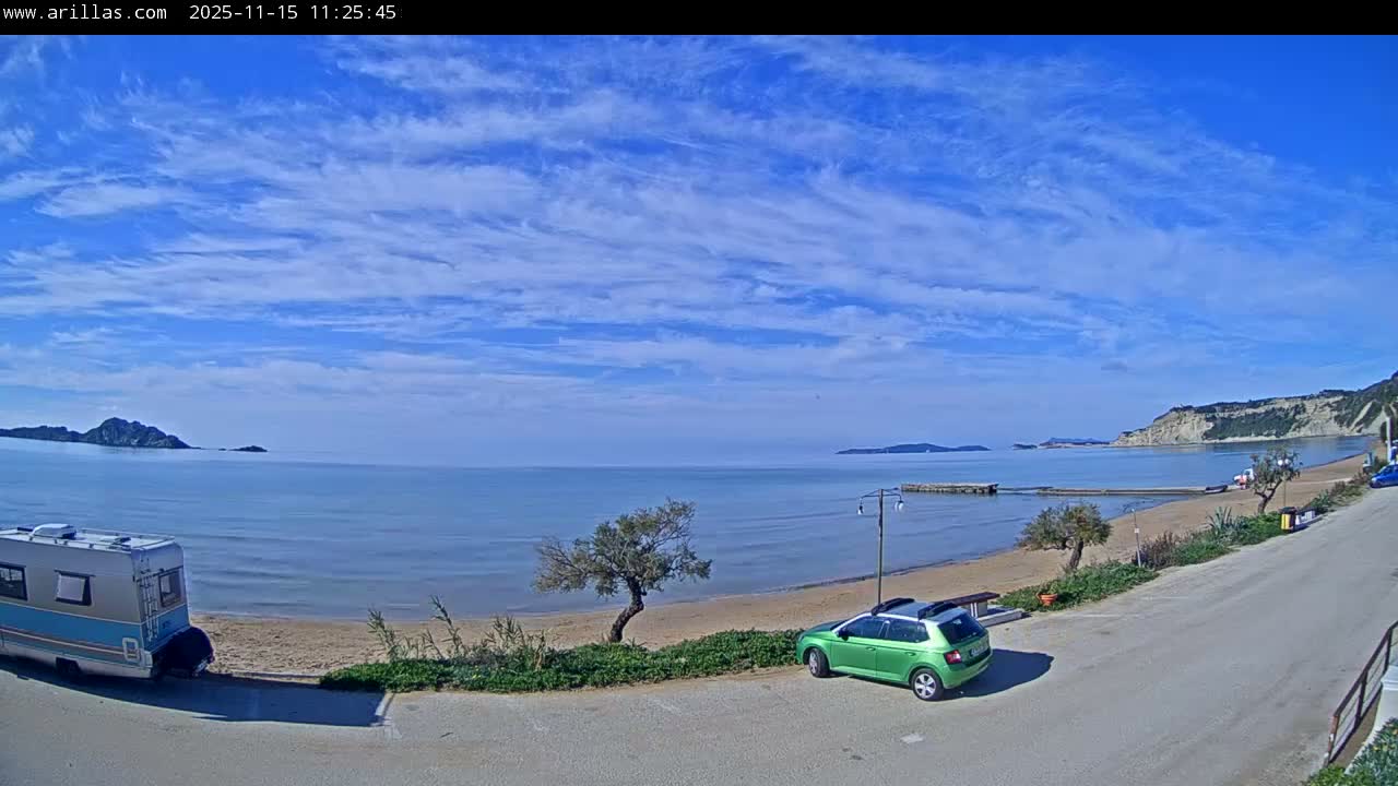 A sunny coastal scene features a sandy beach bordered by calm blue sea with distant islands, a camper van parked on the sand, and a green car on a nearby road, all beneath a bright blue sky dotted with white clouds.