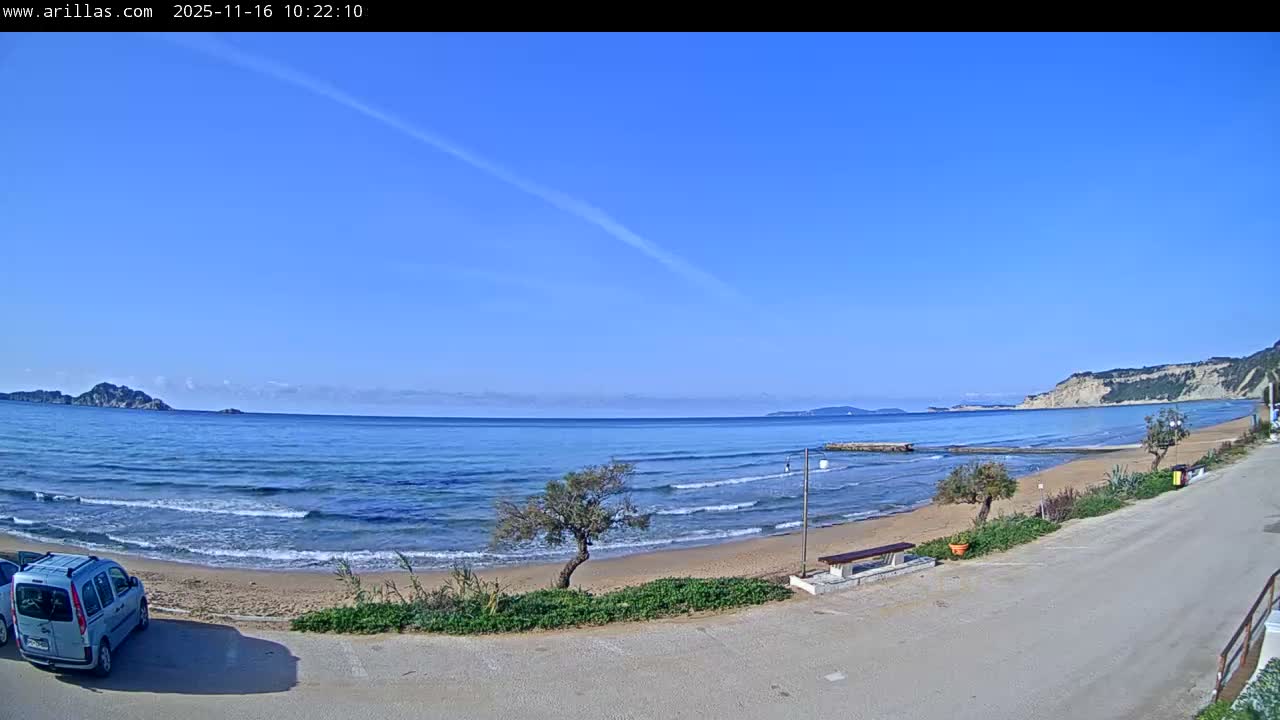 On a clear sunny day, a light blue van is parked on a paved area overlooking a sandy beach with gentle waves, stretching towards distant islands on the left and a prominent cliff face on the right, all under a bright blue sky.