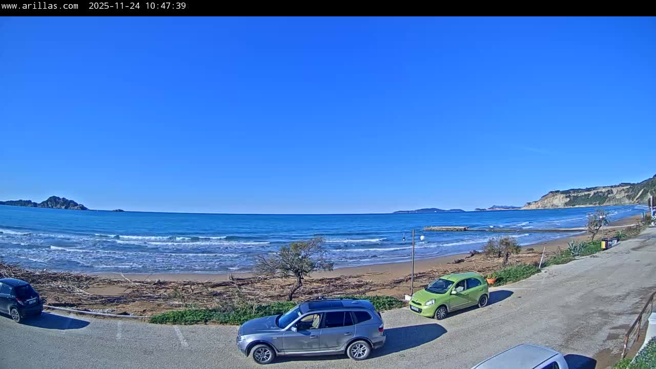 A sunny, clear blue sky overlooks a wide beach with crashing waves and driftwood, distant islands and cliffs, and several cars parked on a paved area in the foreground.