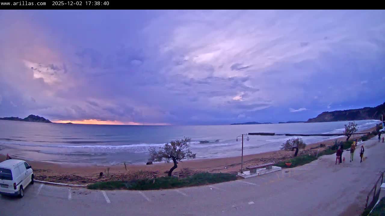 A dramatic cloudy evening over a coastal scene features a sandy beach and choppy ocean waters under a sky painted in vibrant purples, oranges, and blues, with distant hills, a parked white van, and several people walking along a paved path.