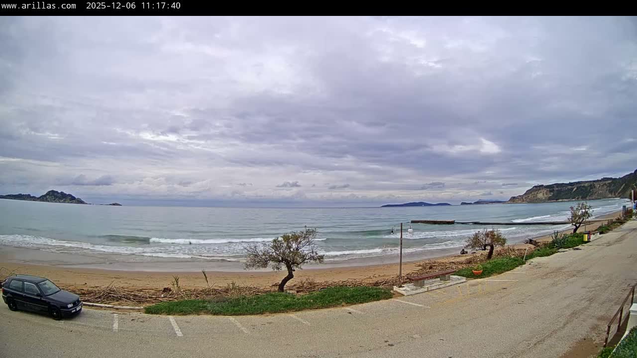 A wide coastal view under a heavily overcast sky depicts a quiet beach with gentle waves, a black car parked on a sandy lot, a partial wooden pier or breakwater in the water, and distant mountainous coastlines and islands.