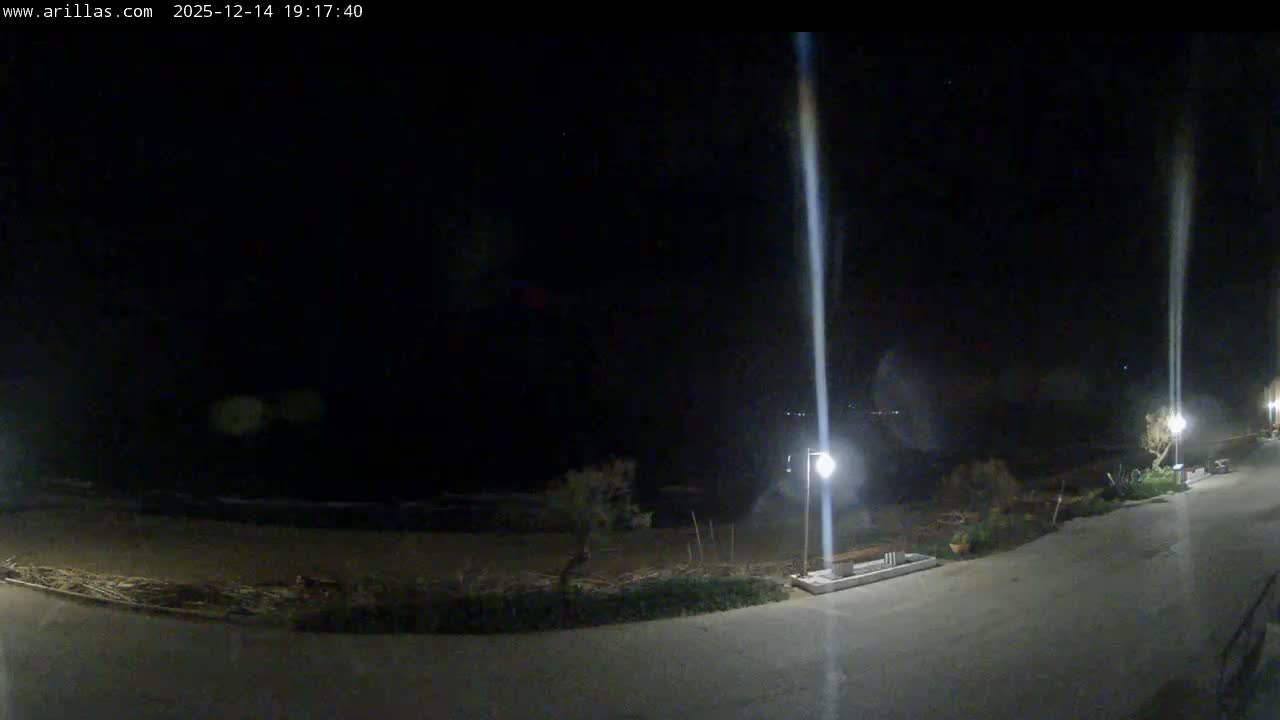 A wide coastal view under a heavily overcast sky depicts a quiet beach with gentle waves, a black car parked on a sandy lot, a partial wooden pier or breakwater in the water, and distant mountainous coastlines and islands.