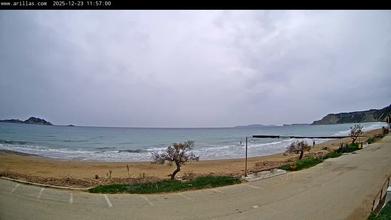 The image displays an overcast day at a wide sandy beach with turquoise waves breaking on the shore, a paved area with a bench in the foreground, and distant hills and small islands under a cloudy sky.