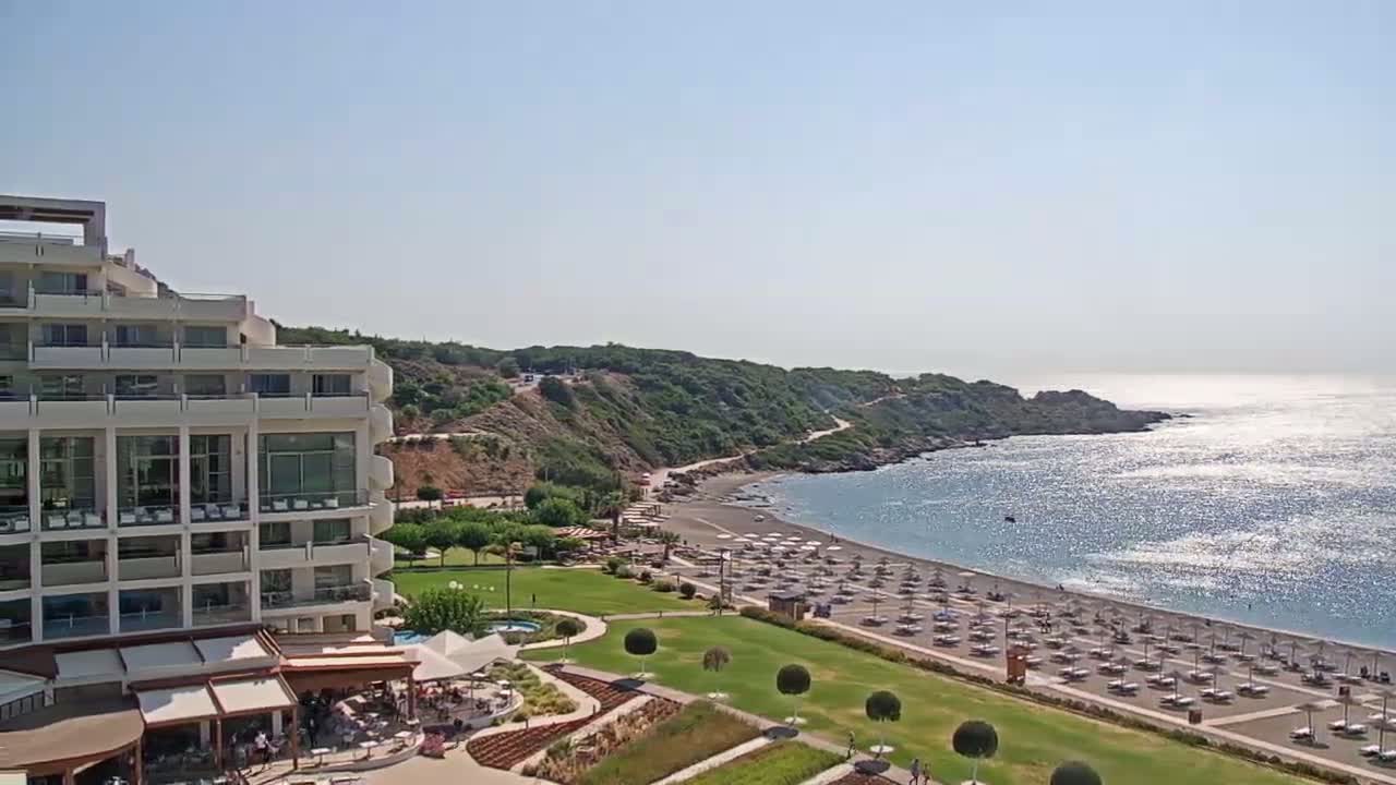 A large, multi-story white hotel sits beside a sandy beach with numerous umbrellas and lounge chairs, overlooking a calm, sunlit ocean on a clear day.
