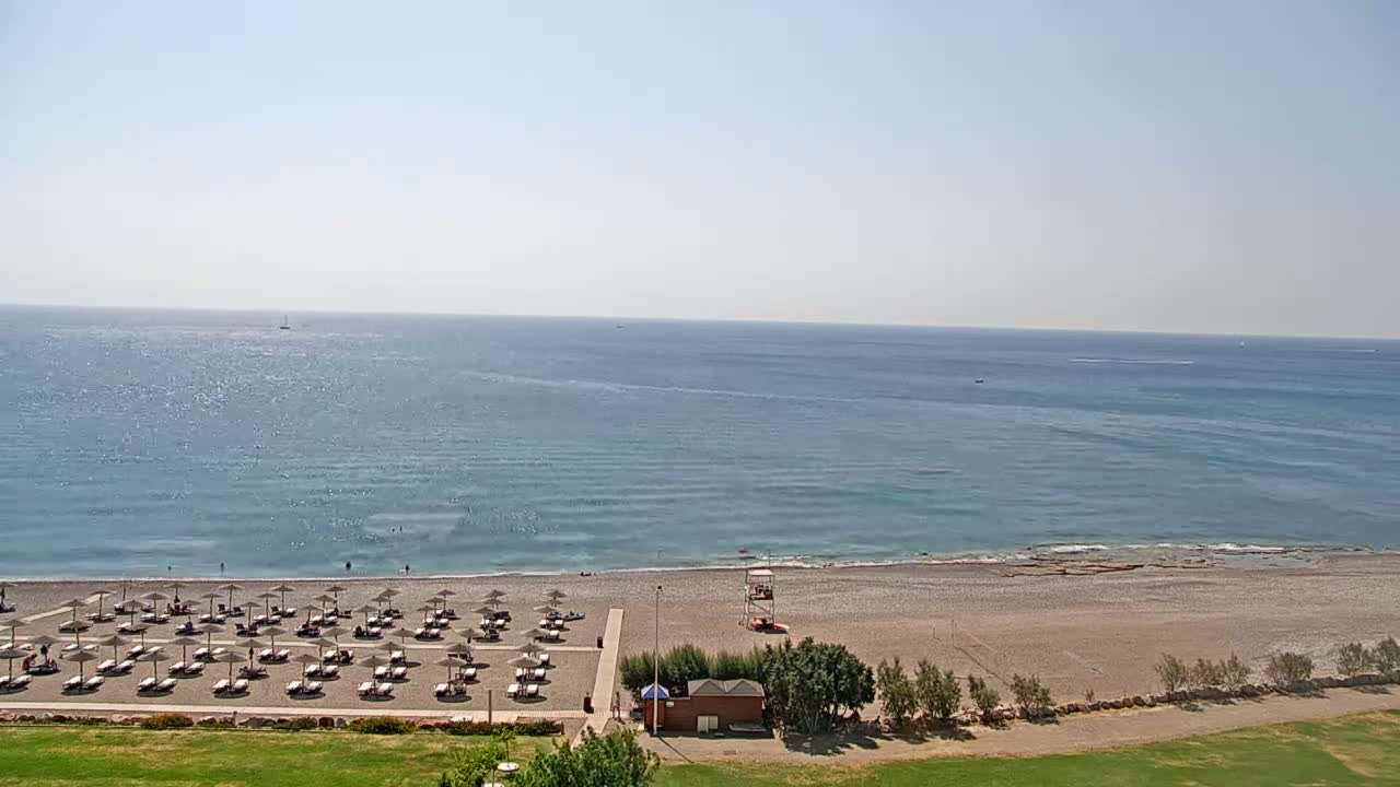 A calm, sunny day reveals a pebble beach with rows of umbrellas and lounge chairs, a lifeguard stand, and a tranquil blue sea stretching to the horizon.