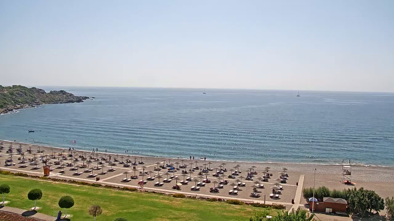 A beach with many umbrellas and lounge chairs on a pebble shore, next to calm blue water under a clear sky.