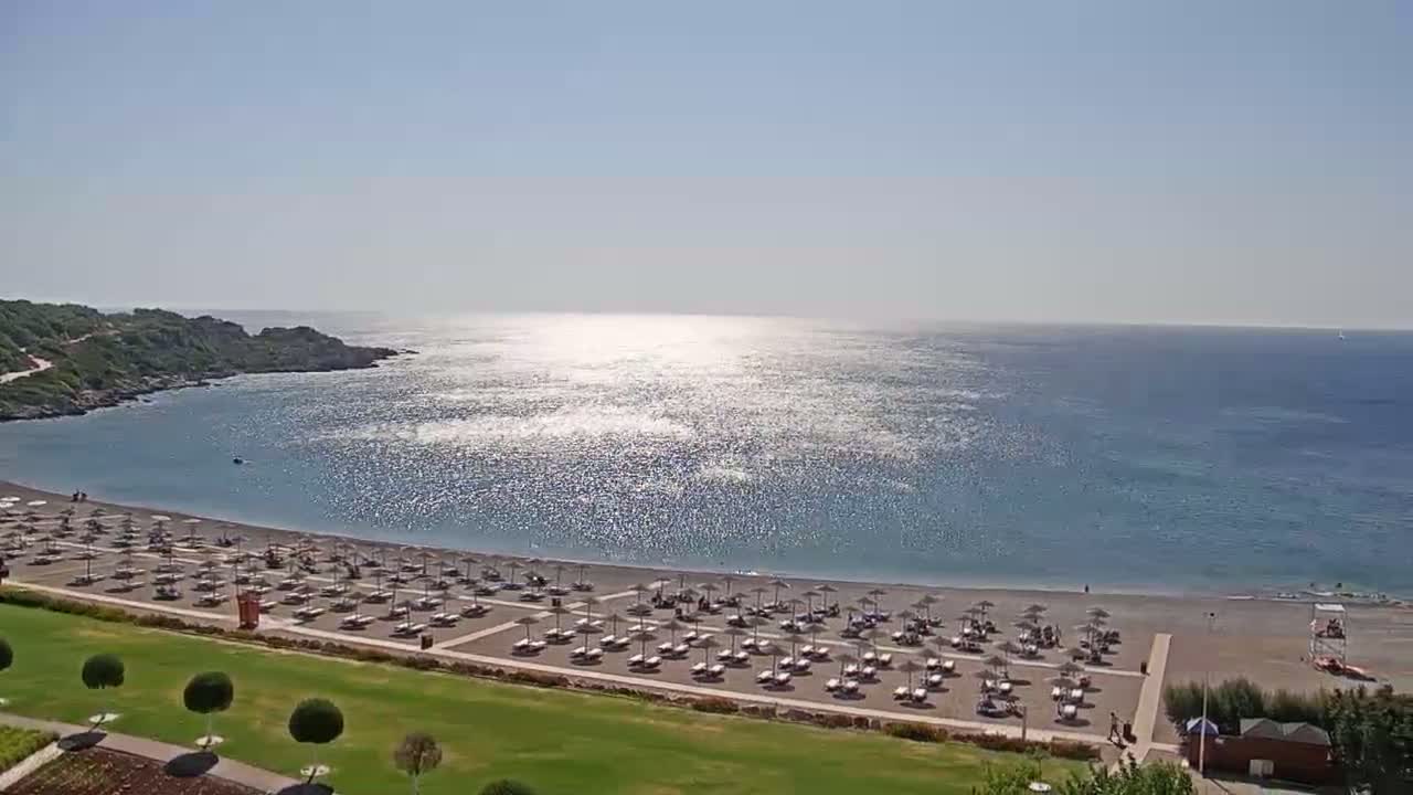 A beach with numerous umbrellas and lounge chairs sits on a calm, sun-dappled sea next to green lawns and low hills under a clear sky.