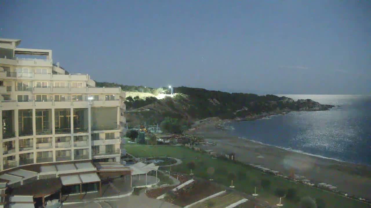 The image displays a coastal resort at night, featuring a large, brightly lit hotel on the left, a landscaped lawn and beach in the middle, and a dark, tree-covered peninsula extending into a shimmering sea under a clear, dark blue sky.