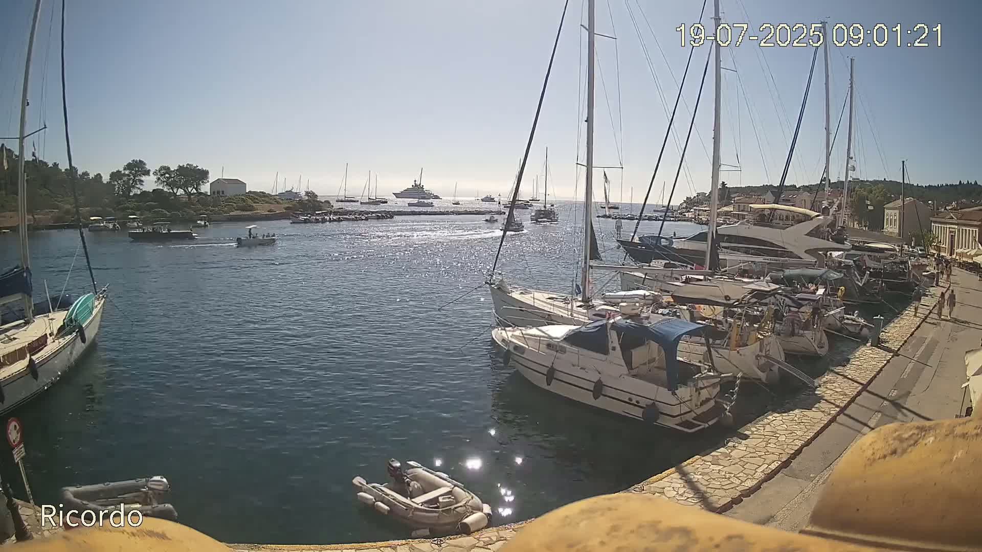 Numerous sailboats and motorboats are docked in a harbor on a sunny day, with additional vessels visible in the distance.