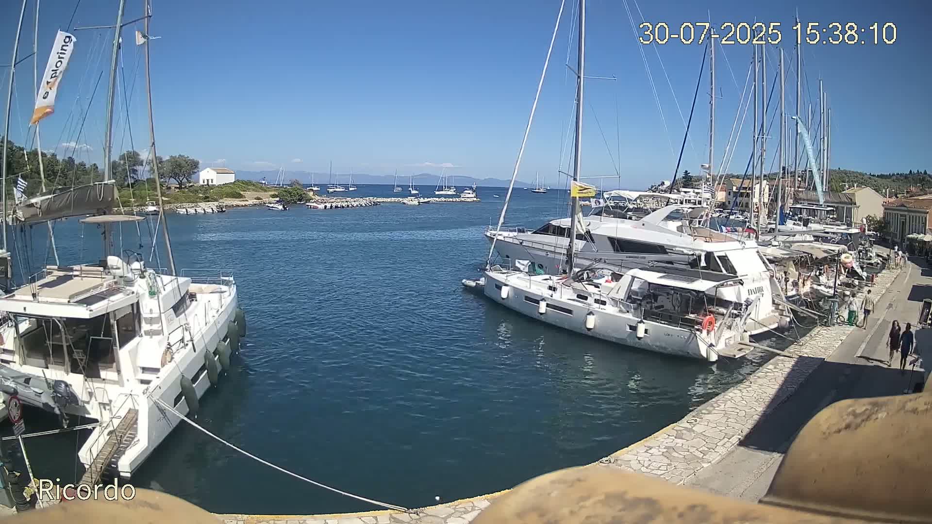 Numerous sailboats and motorboats are docked in a harbor on a sunny day, with additional vessels visible in the distance.