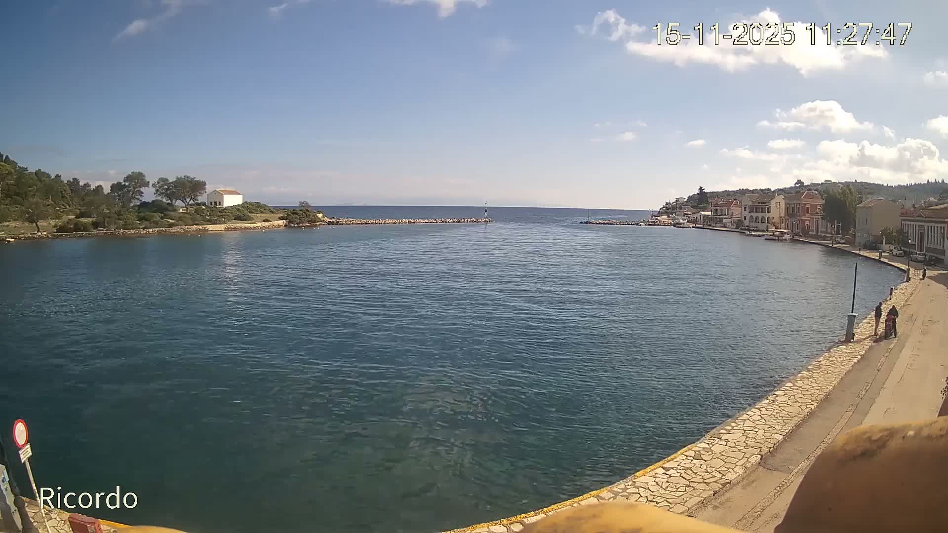 A vibrant coastal town with colorful buildings lines a calm bay under a sunny sky with scattered clouds, opposite a tree-covered peninsula featuring a white building, all leading to the open sea marked by a small lighthouse.