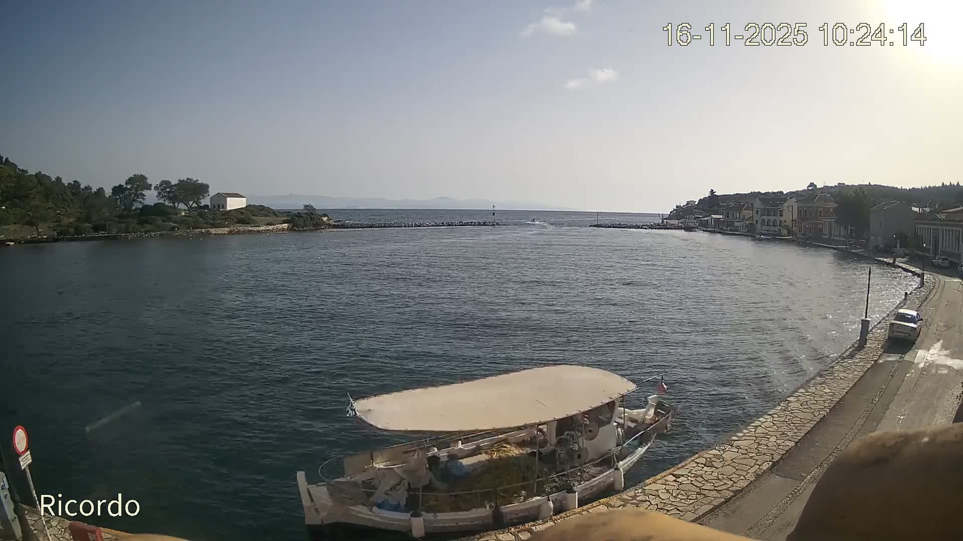 A sunny outdoor scene depicts a calm bay where a white fishing boat is moored at a stone quay bordering a coastal town, with a tree-covered island featuring a white building opposite, and the open sea visible through an entrance flanked by breakwaters.