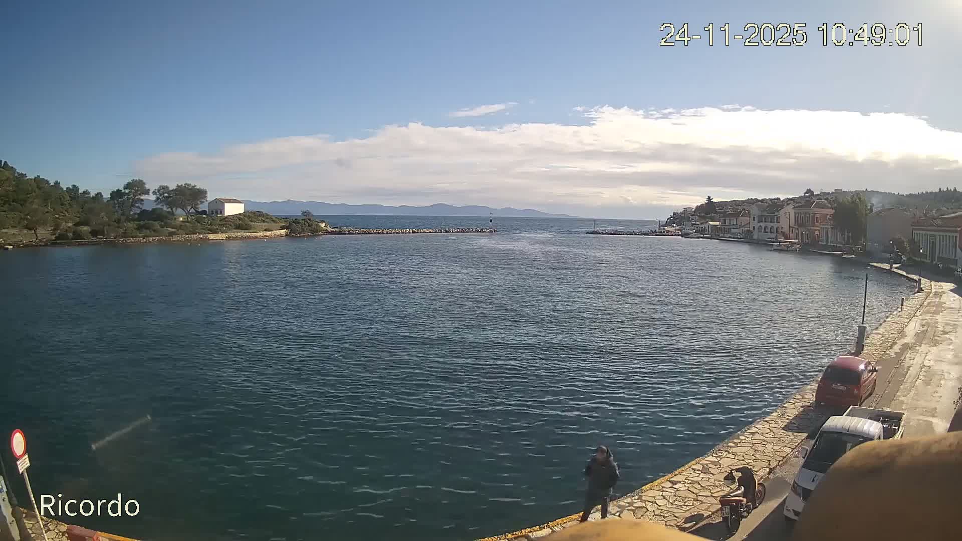 The image depicts a calm bay with deep blue water, framed by a wooded peninsula featuring a small white building on the left, and a charming waterfront town with buildings lining a paved promenade on the right, all under a bright, partly cloudy sky.