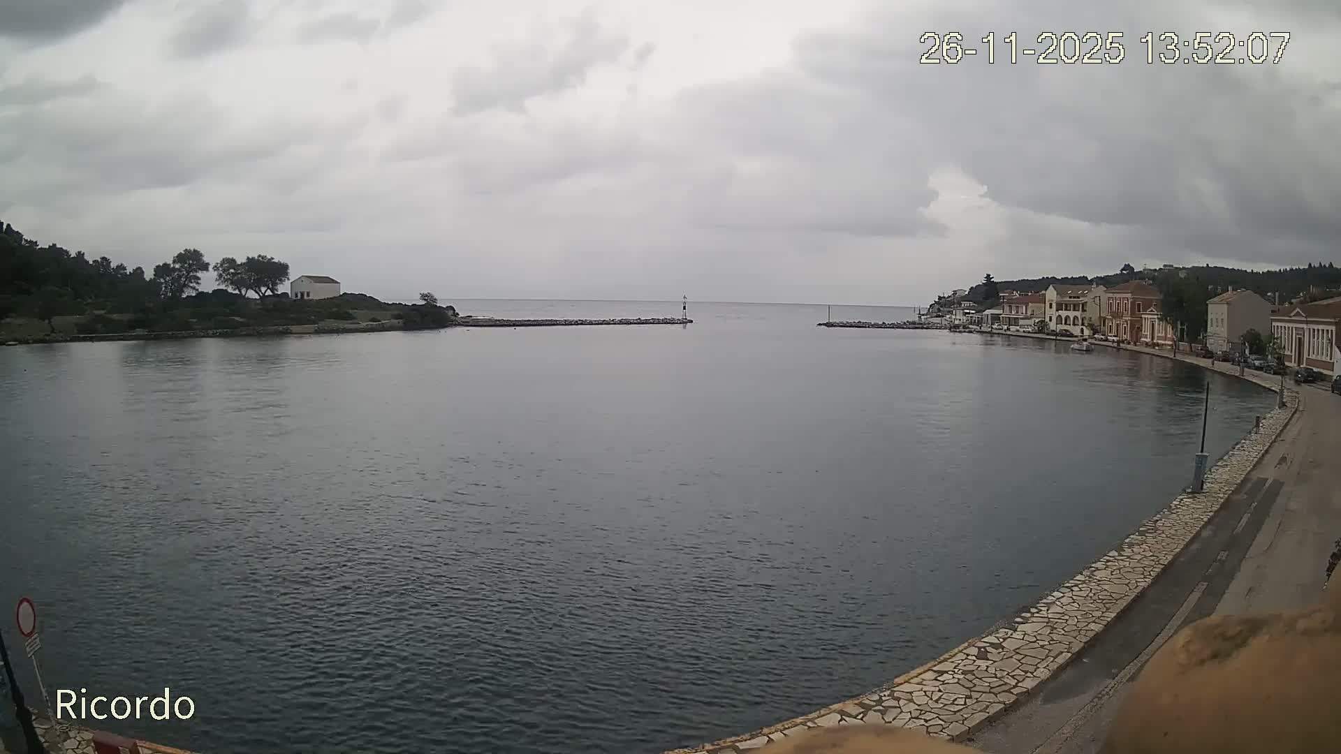 The image depicts a calm bay with deep blue water, framed by a wooded peninsula featuring a small white building on the left, and a charming waterfront town with buildings lining a paved promenade on the right, all under a bright, partly cloudy sky.