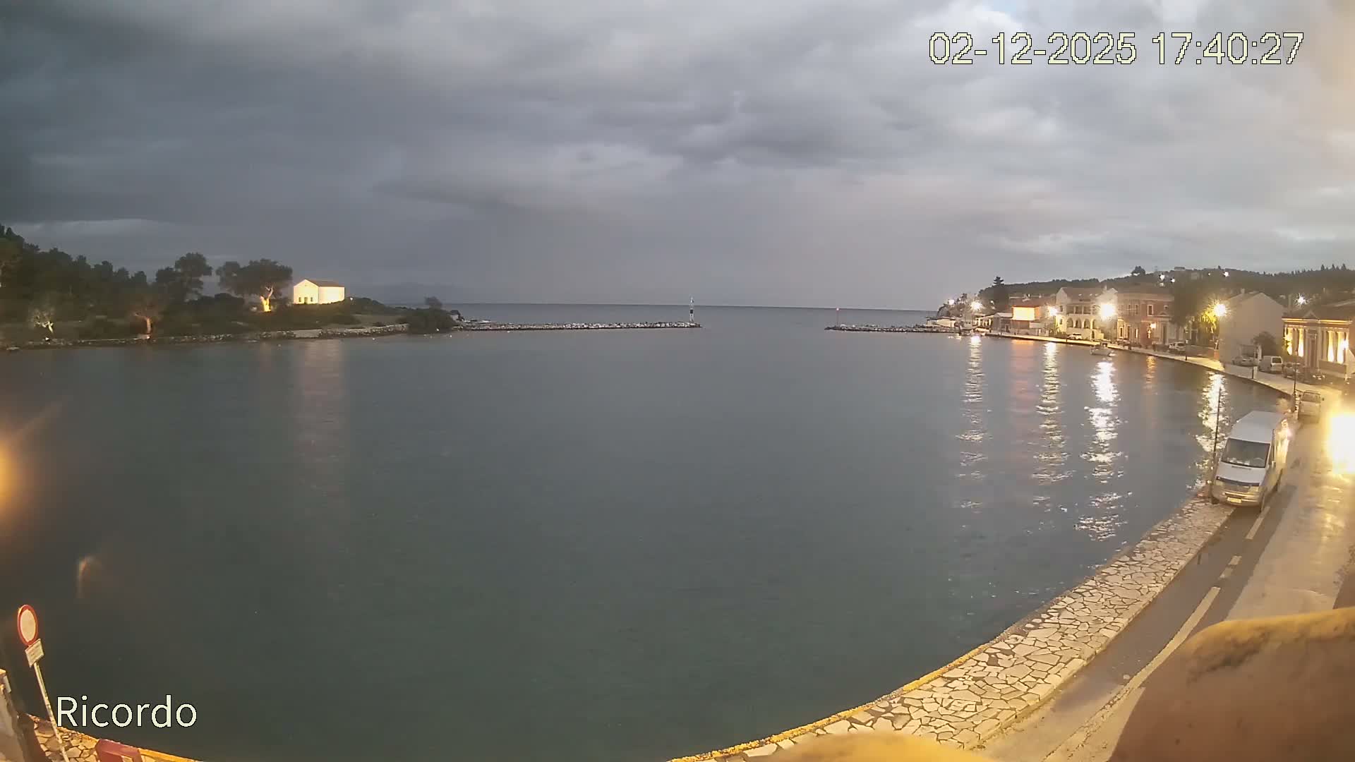 An overcast and rainy evening view of a coastal town's harbor shows illuminated buildings reflecting in the dark water and a wet promenade, with a distant lighthouse and a bright building on a peninsula.