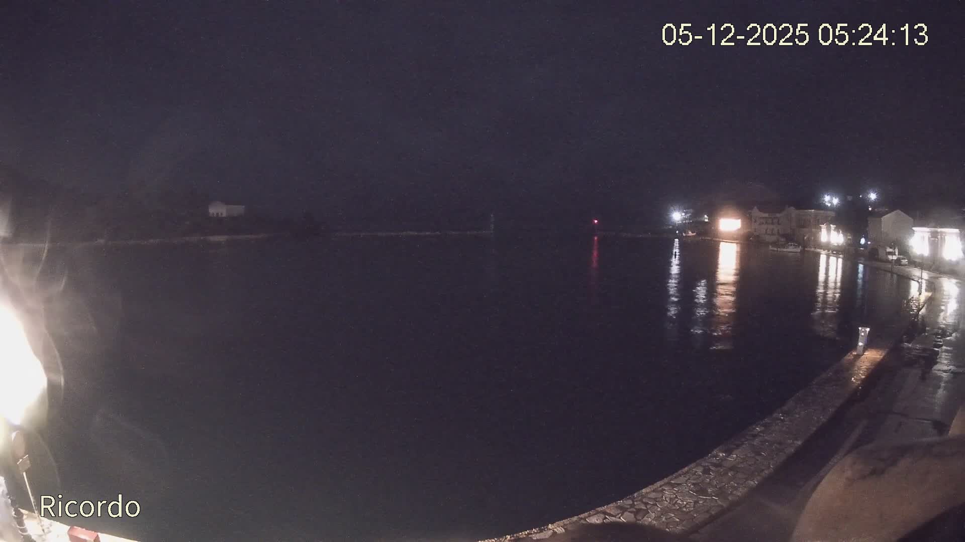 An overcast and rainy evening view of a coastal town's harbor shows illuminated buildings reflecting in the dark water and a wet promenade, with a distant lighthouse and a bright building on a peninsula.