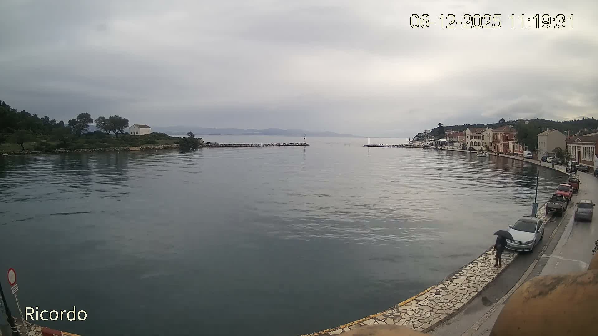 An overcast and rainy day is depicted over a tranquil coastal bay, featuring historic-looking buildings lining the waterfront, a distant point with a white building, and a person walking with an umbrella on a wet promenade.