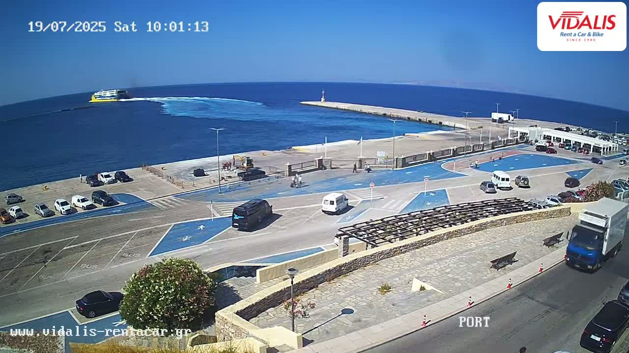 A ferry departs from a harbor on a sunny day, with cars parked in the surrounding lot.