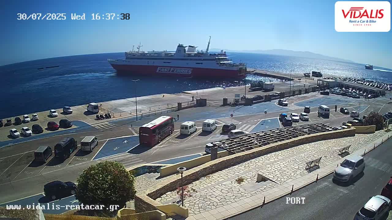 A ferry departs from a harbor on a sunny day, with cars parked in the surrounding lot.