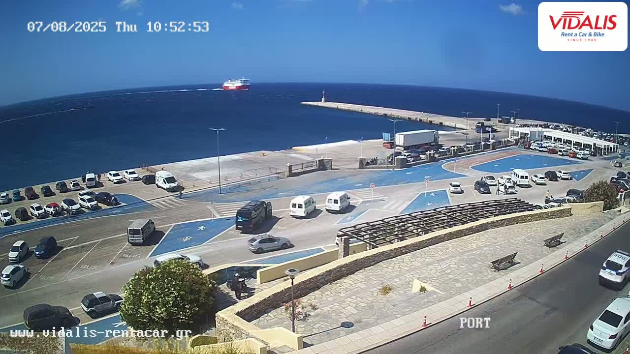 A ferry approaches a port on a sunny day, where numerous cars and vans are parked in a lot, with a few more driving on the roads surrounding the area.