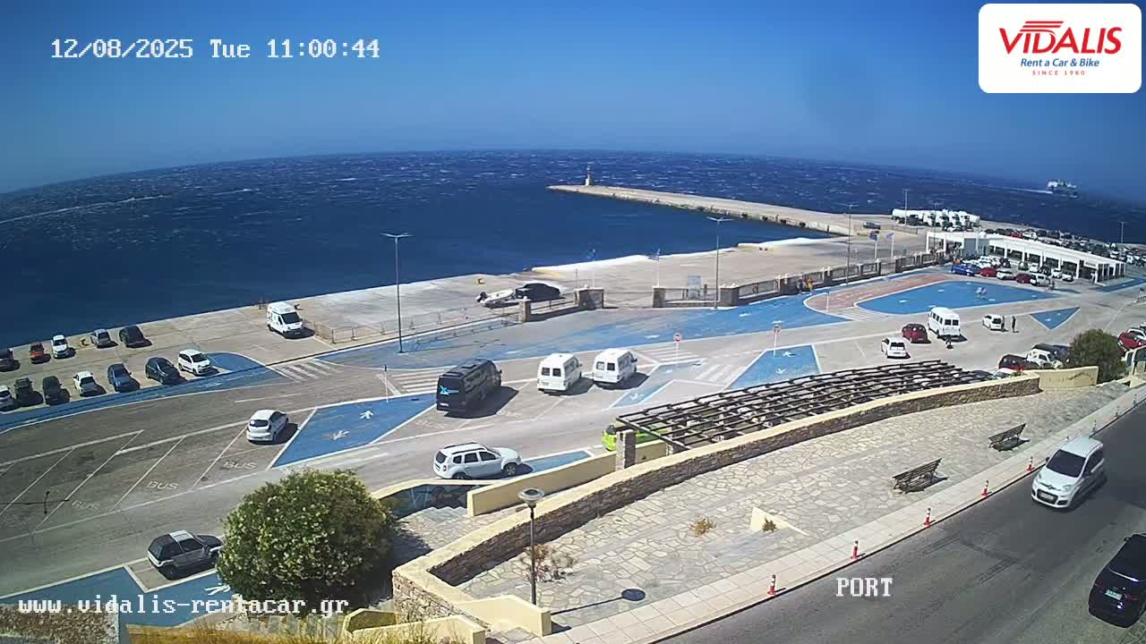 A seaside port with a parking lot and several vehicles, viewed on a sunny day with a windy sea.