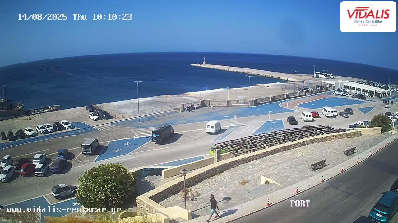A sunny day overlooking a harbor with cars parked in a lot, a person walking by, and a calm blue sea extending to the horizon.