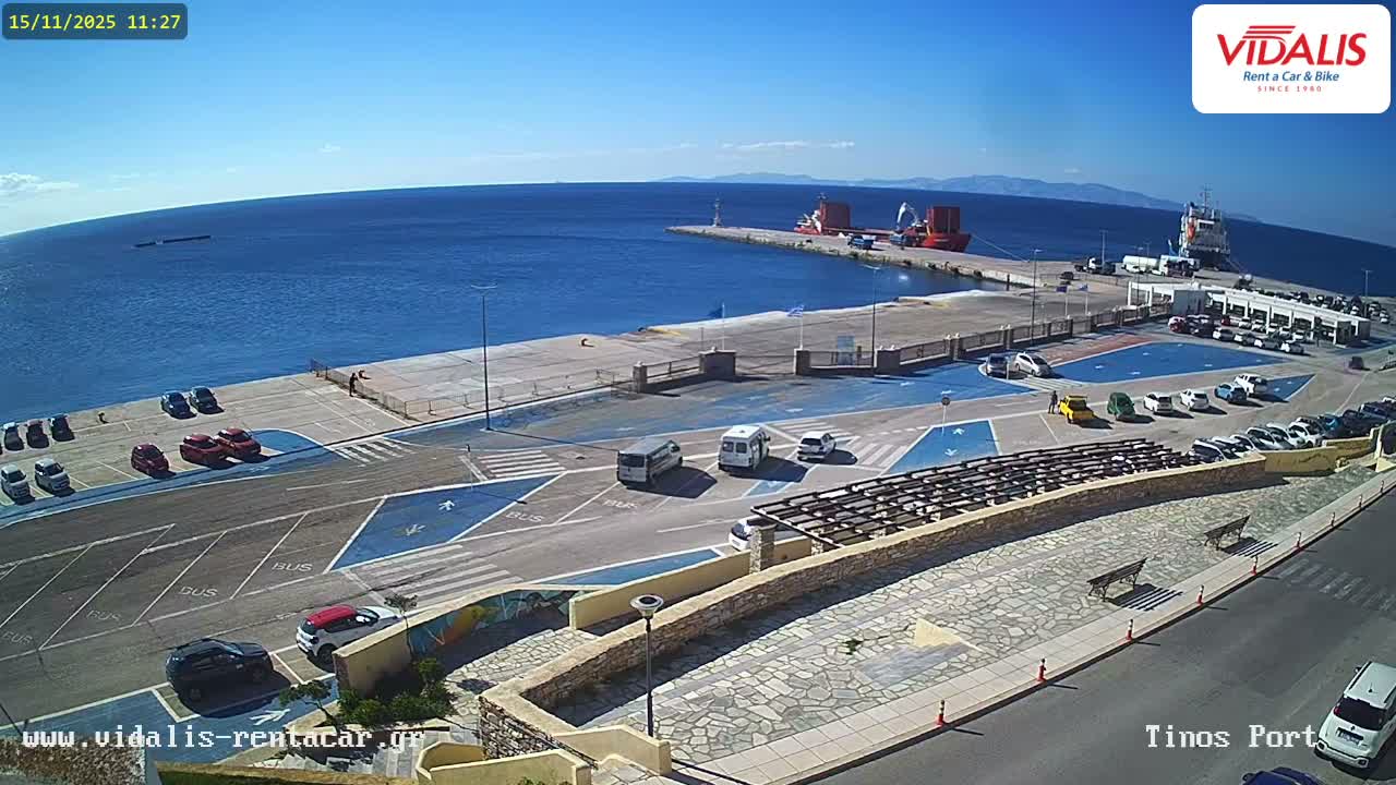 This wide-angle view captures a sun-drenched port under a clear blue sky, showcasing deep blue water with docked ships, extensive paved areas filled with parked cars and moving vehicles including buses and vans, along with pedestrian walkways and distant mountains.