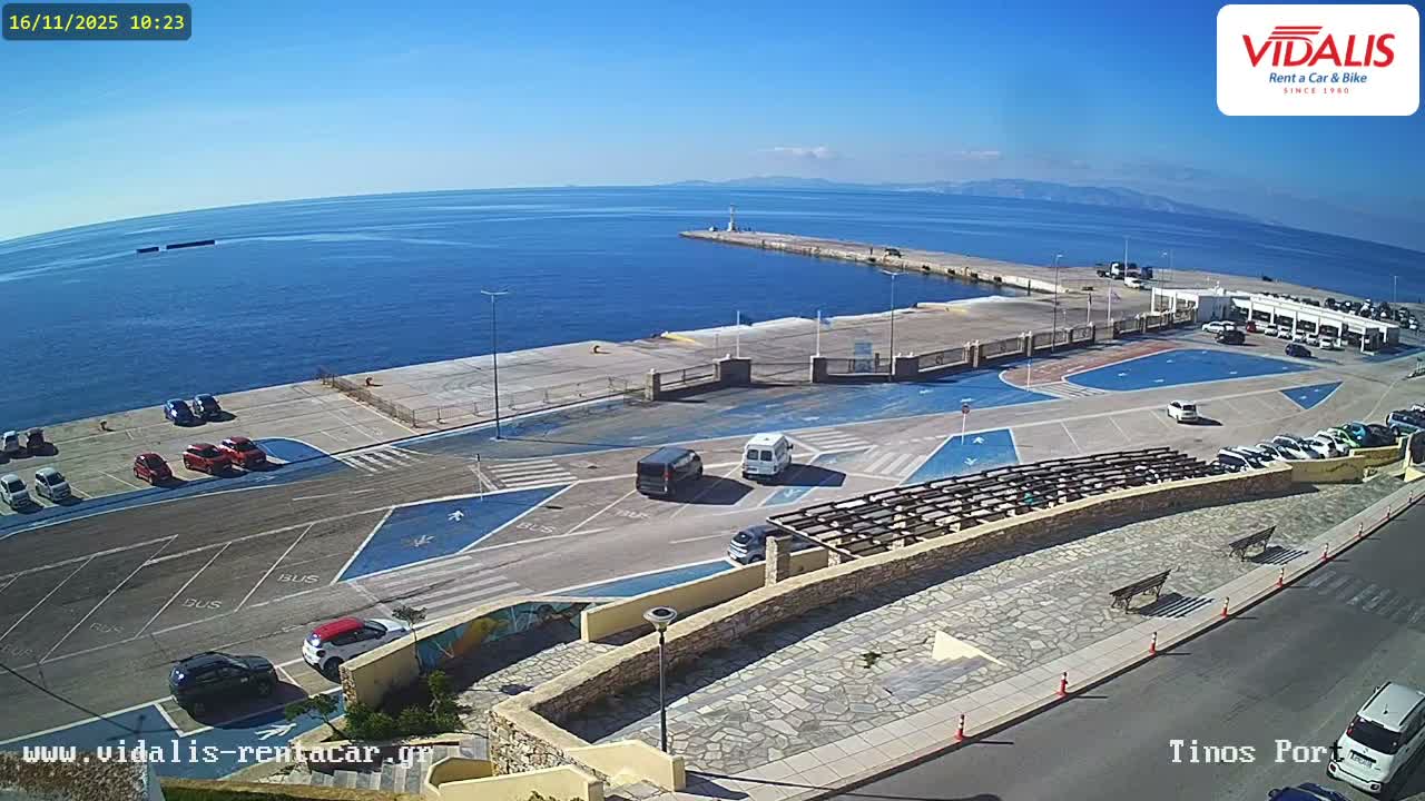 On a bright, clear, and sunny day, a port terminal with various cars parked on a paved lot sits beside a calm blue sea, where a long pier topped with a lighthouse extends into the water towards distant land.